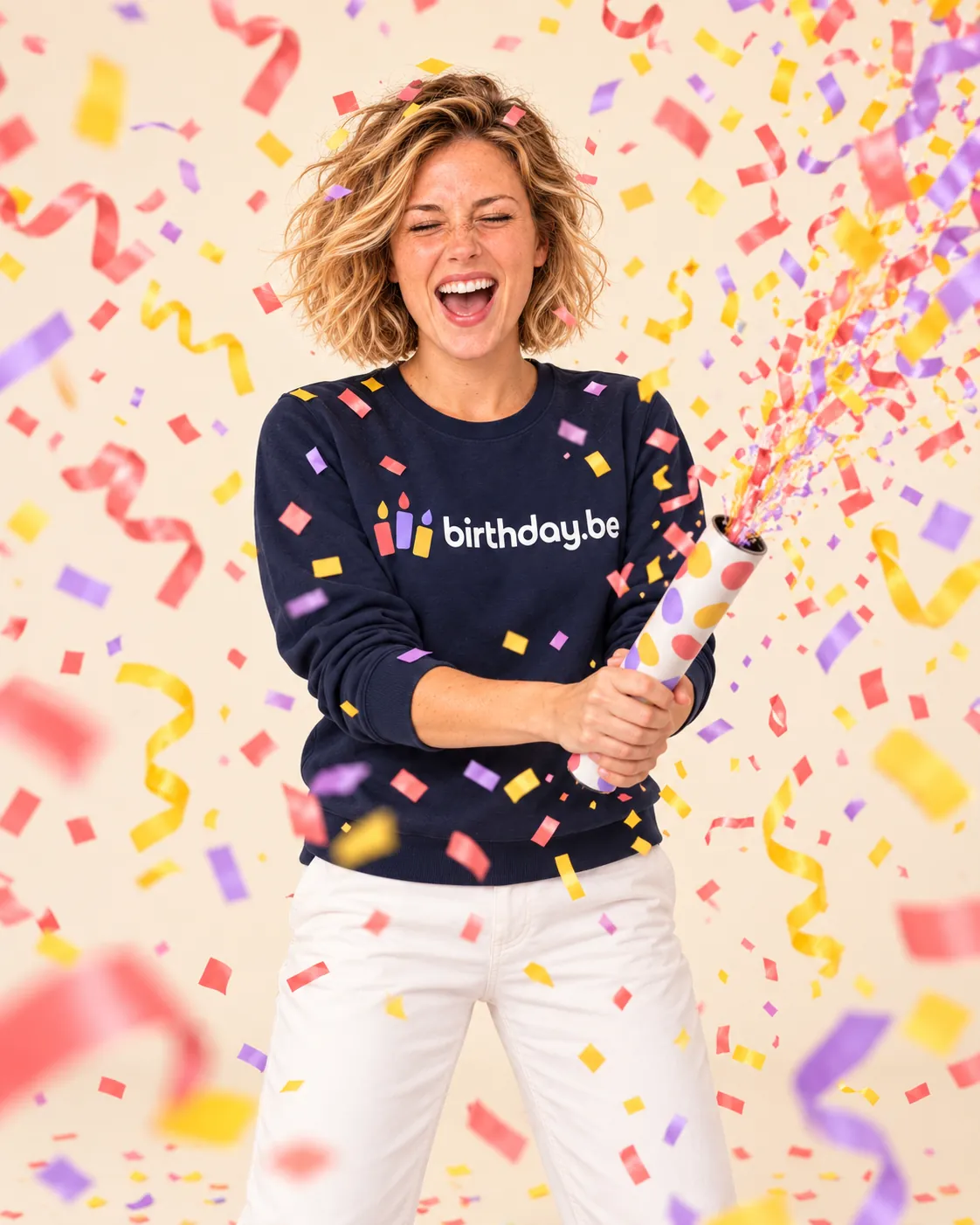 Lachende persoon in een donkerblauwe sweater met birthday.be logo, omgeven door vallende gekleurde confetti en serpentines op een crème achtergrond