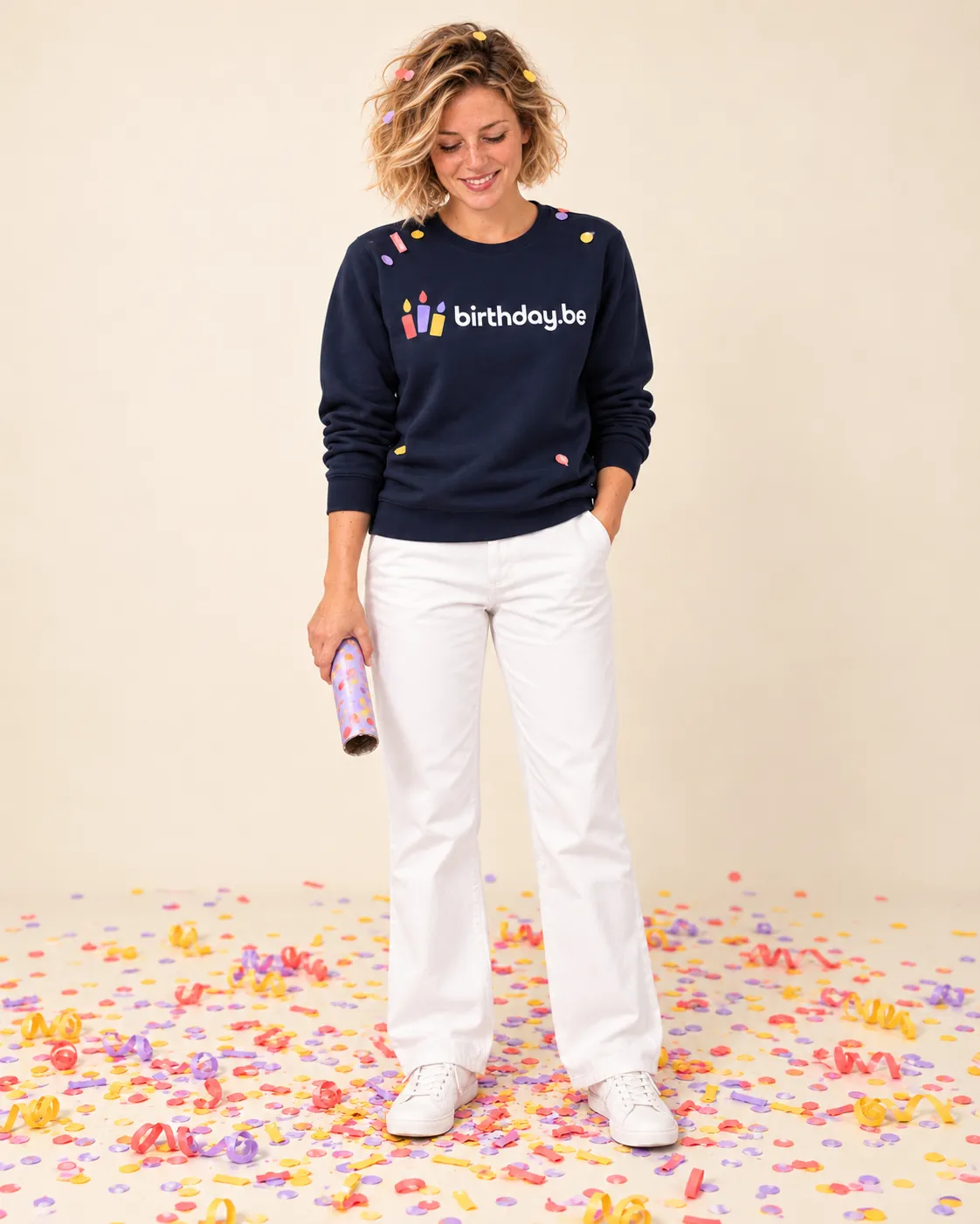 Persoon draagt donkerblauwe sweatshirt met birthday.be logo en witte broek, omgeven door kleurrijk confetti
