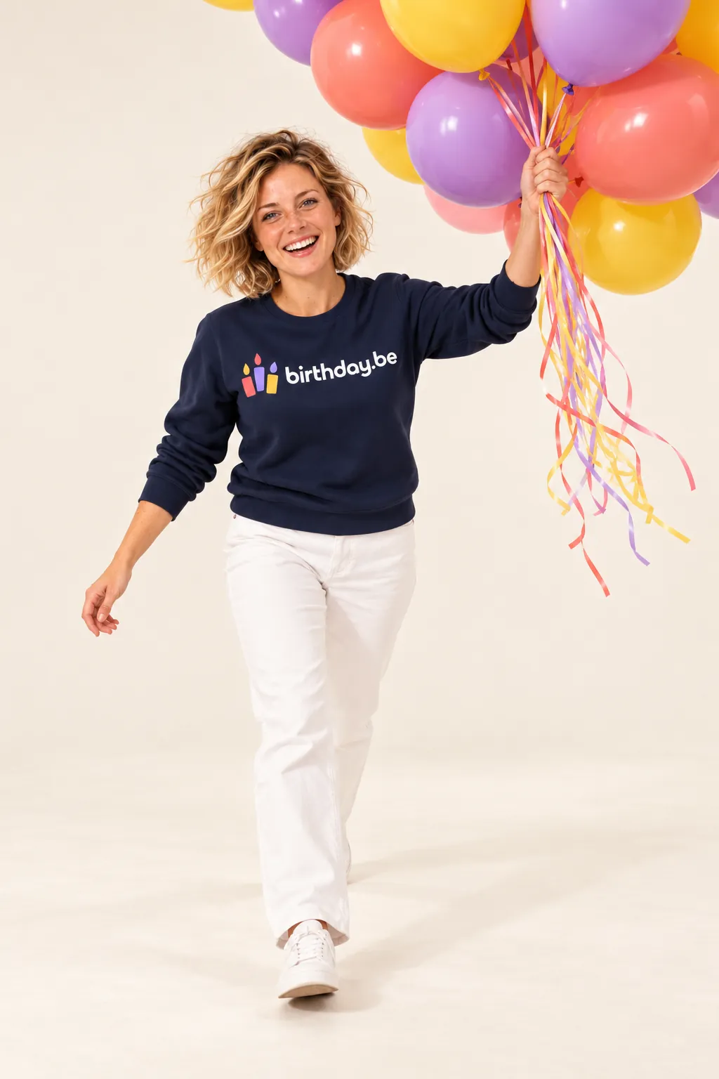 Fien in donkerblauwe sweater met birthday.be logo houdt een bos kleurrijke ballonnen vast tegen een lichte achtergrond