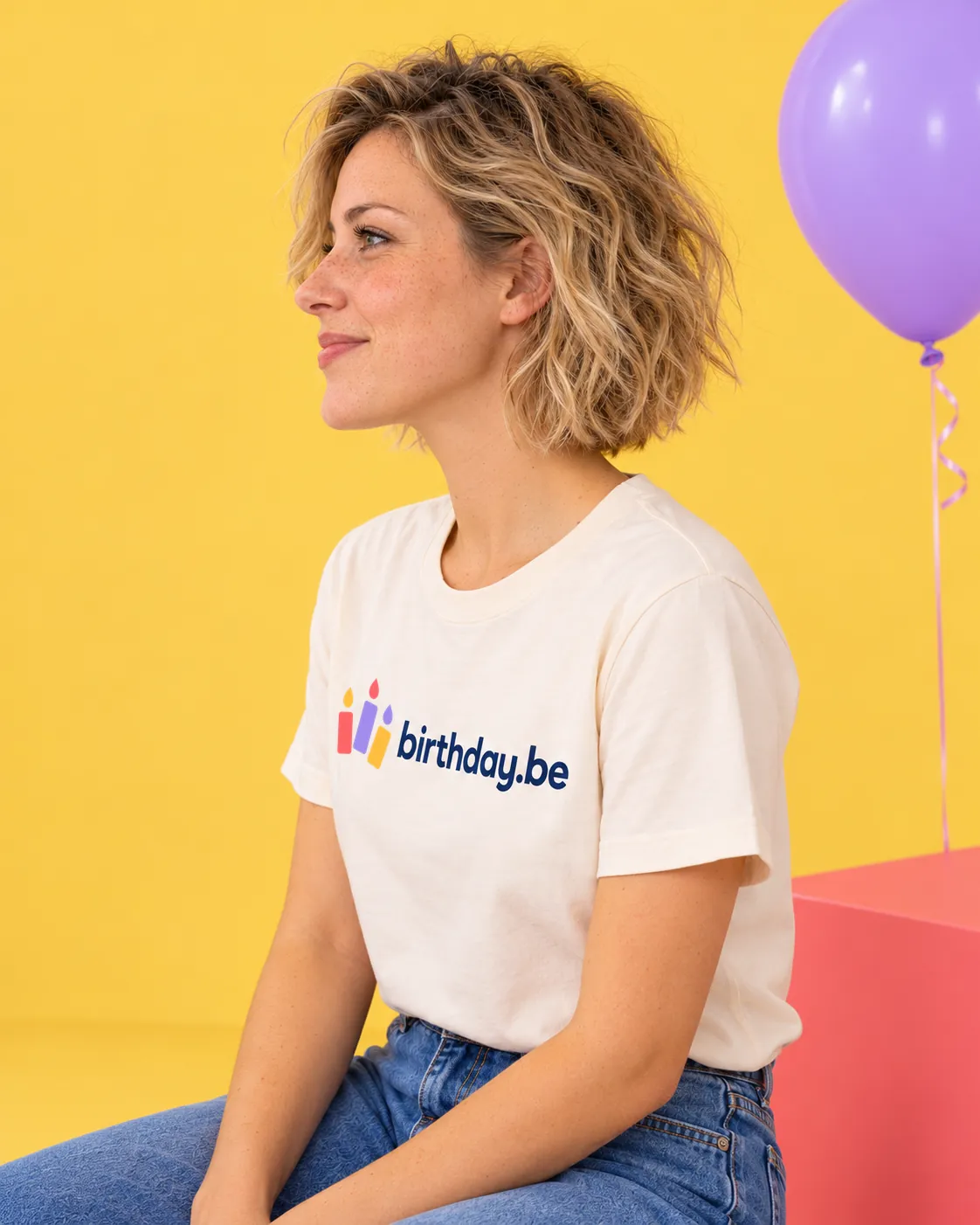 Persoon draagt een wit t-shirt met het logo van birthday.be tegen een gele achtergrond met een paarse ballon