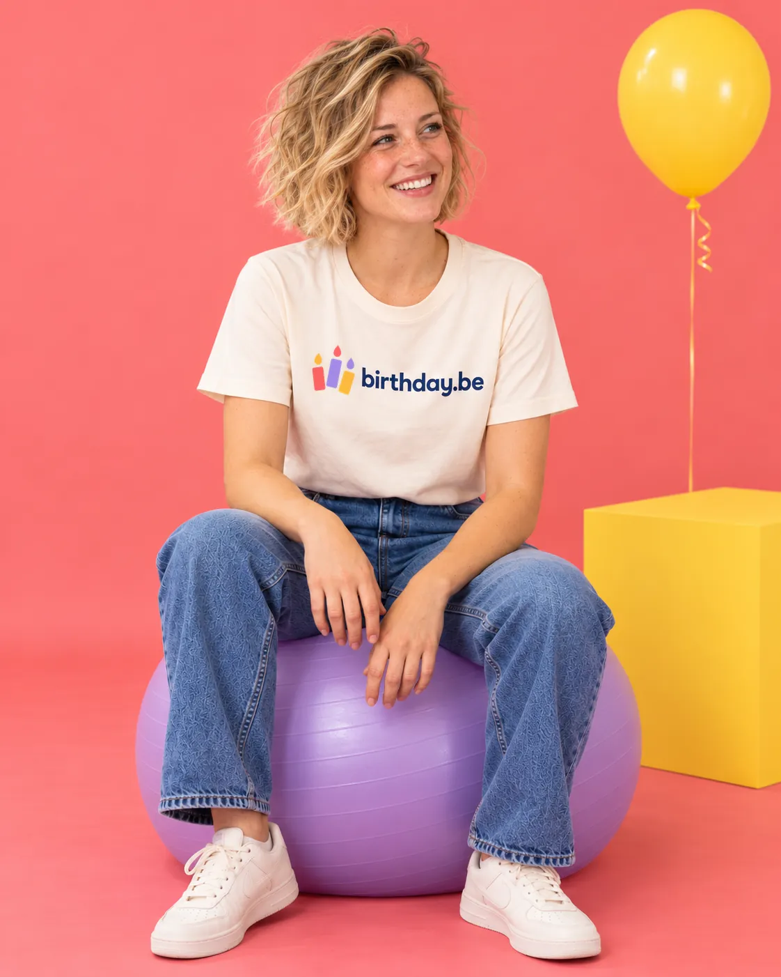 Persoon zit lachend op een paarse bal tegen een roze achtergrond, draagt een birthday.be t-shirt en jeans, met een gele ballon op de achtergrond